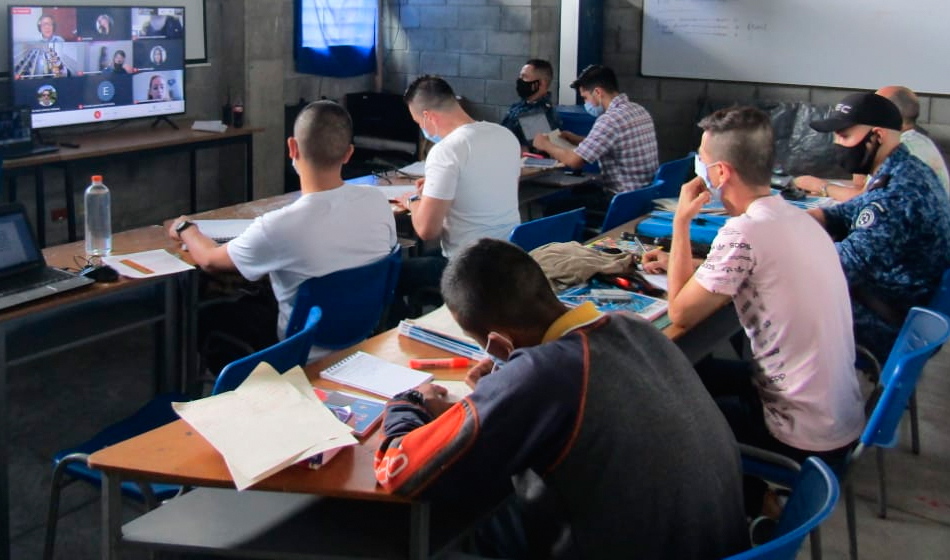 Población carcelaria tomando clases de comunicación social hombres en aula de clase observando videoconferencia en computador.