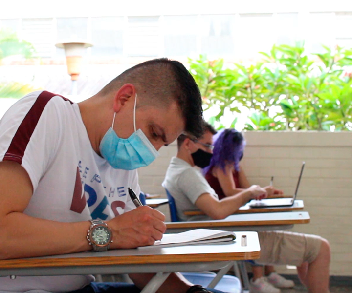 Piloto para el regreso a clase en Pereira Estudiante usa tapaboca y escribe en silla de universidad. Al fondo se ven dos compañeros suños escuchando la clase.