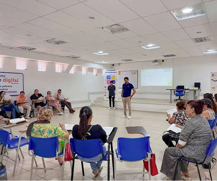 En un aula amplia e iluminada, un hombre con camisa azul y pantalón beige se encuentra de pie en el centro, hablando con los asistentes. A su lado, otro hombre con ropa oscura observa. Alrededor, varias personas, en su mayoría mujeres, están sentadas en sillas azules con escritorios adjuntos, tomando notas y prestando atención. En el fondo, hay una pantalla proyectando información, indicando un entorno educativo