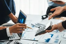 Manos de profesionales revisando gráficos financieros en una laptop y documentos impresos mientras utilizan una calculadora durante una reunión de análisis financiero.