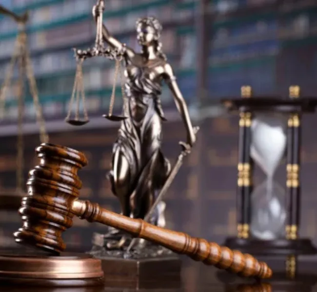 Balanza de la justicia junto a un mazo de juez sobre una mesa, con un reloj de arena y libros de derecho al fondo, representando el concepto de ley, justicia y paso del tiempo en los procesos legales.